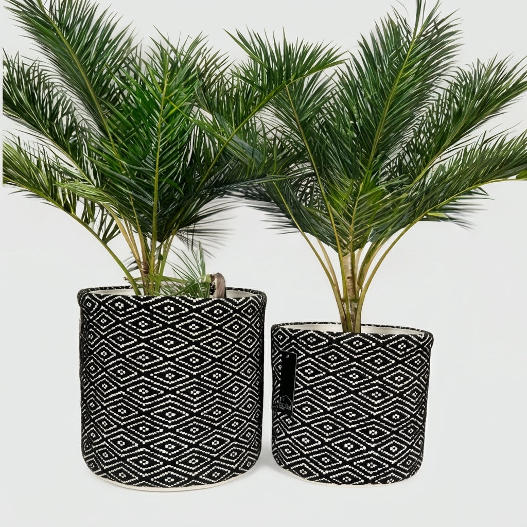 Two potted artificial palm plants in cylindrical black fabric planters with a white geometric diamond pattern, set against a plain white background.