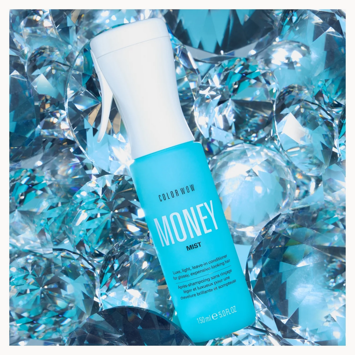 A bottle of Colour Wow Money Mist spray, featuring a bright turquoise blue body and a white spray nozzle, nestled among reflective, faceted blue and silver crystals.