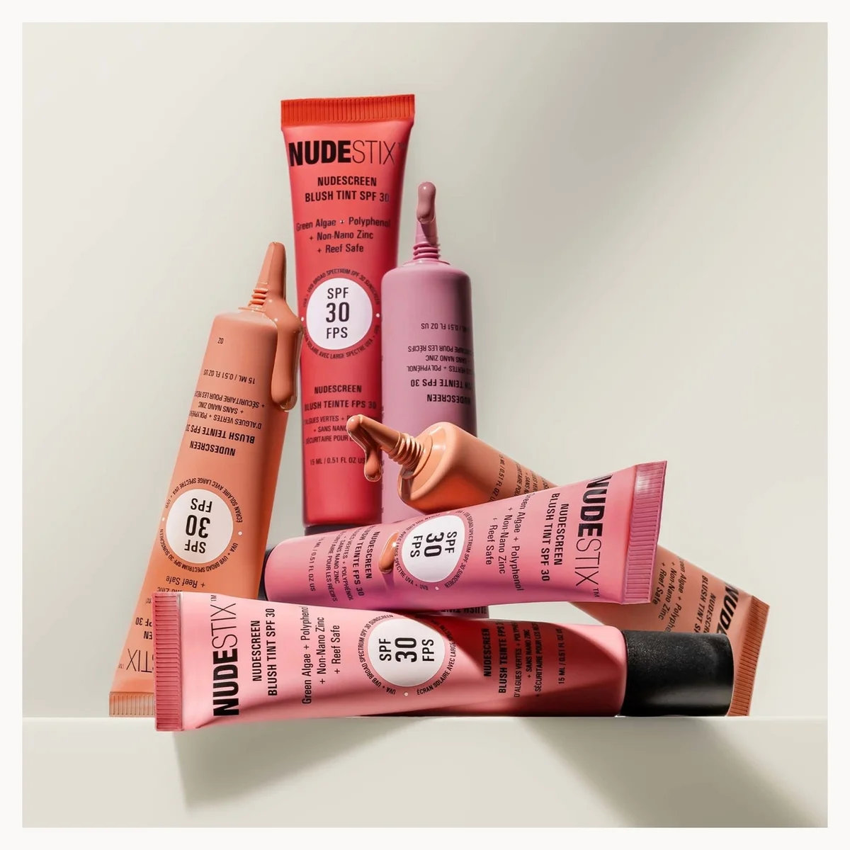 Four tubes of Nudesty moisturizing sunscreen in pink, coral, and mauve shades, arranged on a light beige background.