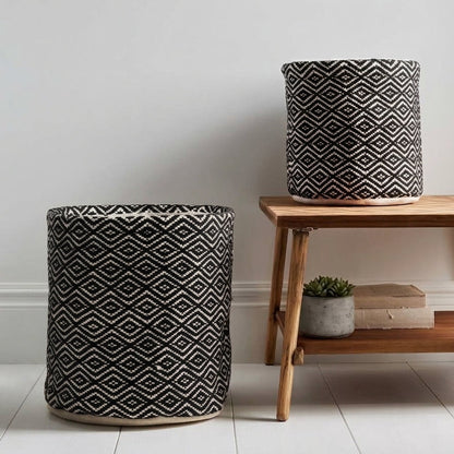 Two cylindrical woven fabric storage bins with a black and white geometric diamond pattern, shown in a styled room setting with a wooden bench and white wall.