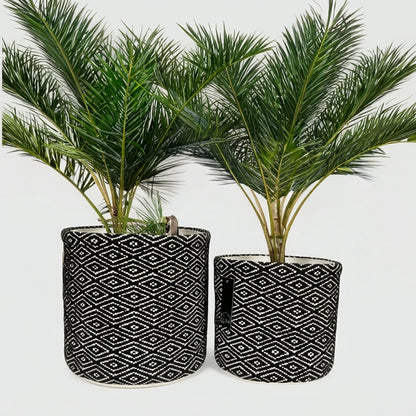 Two potted artificial palm plants in cylindrical black fabric planters with a white geometric diamond pattern, set against a plain white background.