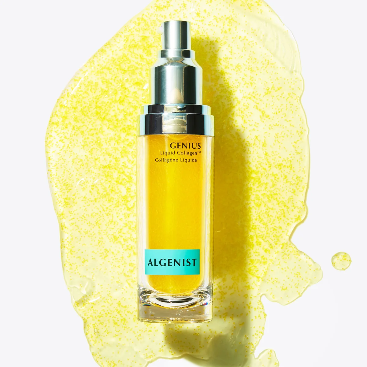 Algenist Genius Liquid Collagen serum in a clear rectangular bottle with a silver pump, filled with golden yellow liquid, set against a bright yellow liquid splash on a white background.