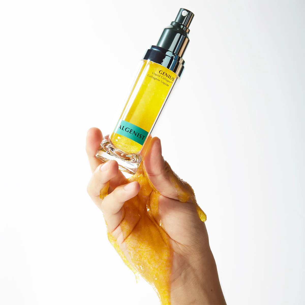 A hand holds a clear bottle of Algenist Genix skincare serum filled with bright yellow liquid, with more yellow gel dripping from the bottle against a clean white background.
