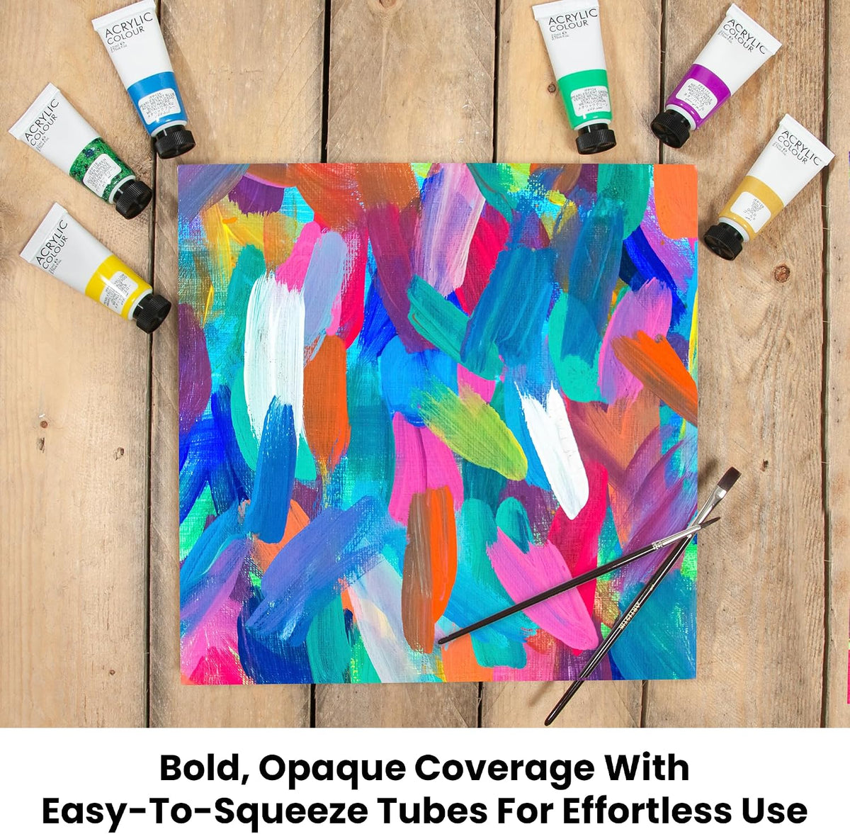 A square canvas with a colorful abstract painted design is laid on a wooden surface, surrounded by multiple squeeze tubes of acrylic paint and two paintbrushes. Text at the bottom reads: 'Bold, Opaque Coverage With Easy-To-Squeeze Tubes For Effortless Use.