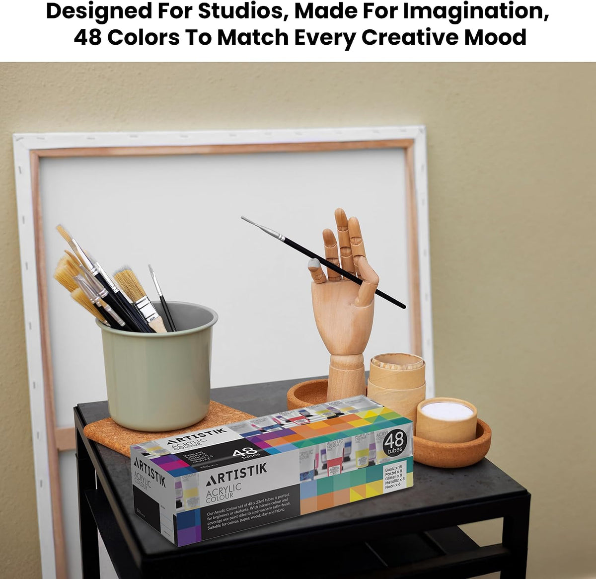 A box of Artistik acrylic paint with 48 colors sits on a black table next to paintbrushes in a cup, a wooden mannequin hand holding a paintbrush, and small wooden cups against a beige wall with a white frame behind it.