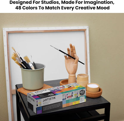 A box of Artistik acrylic paint with 48 colors sits on a black table next to paintbrushes in a cup, a wooden mannequin hand holding a paintbrush, and small wooden cups against a beige wall with a white frame behind it.