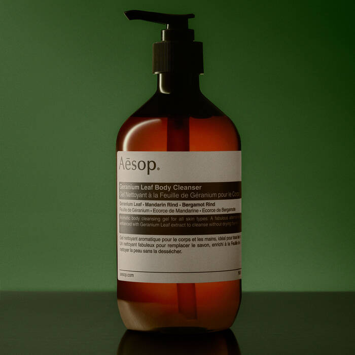 Aesop Geranium Leaf Body Cleanser in an amber brown pump bottle with a white label, set against a dark green background.