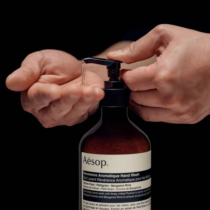 A person's hands dispensing clear hand wash from an amber bottle with a black pump. The bottle has a white label with the brand name Aesop visible, set against a solid black background.