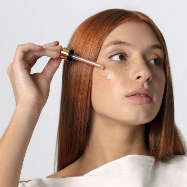 Woman applying dropper of skincare product to her face against a white background