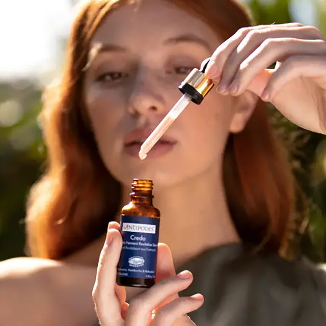 A woman outdoors holds an amber dropper bottle labeled Antifungal Complex while applying a drop of serum from the dropper to her lips.