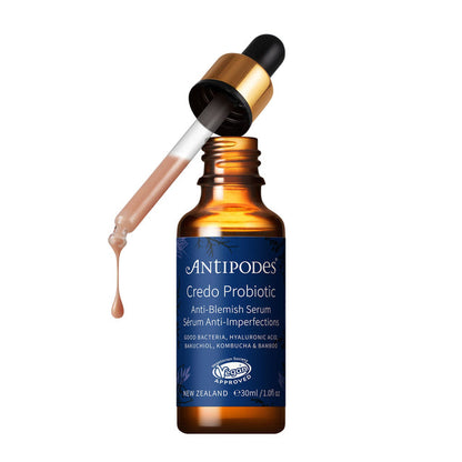 An amber glass dropper bottle of Antipodes Credo Probiotic serum with a dark blue label, shown with the dropper tilted above it and a drop of the serum dripping down on a white background.
