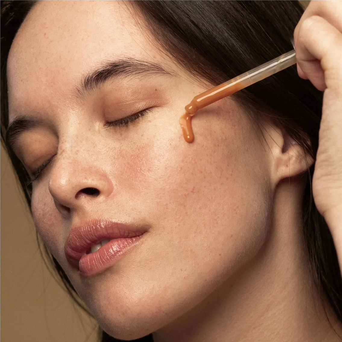 Close-up of a woman's face with a glass dropper applying a warm amber-brown skincare serum to her cheek.