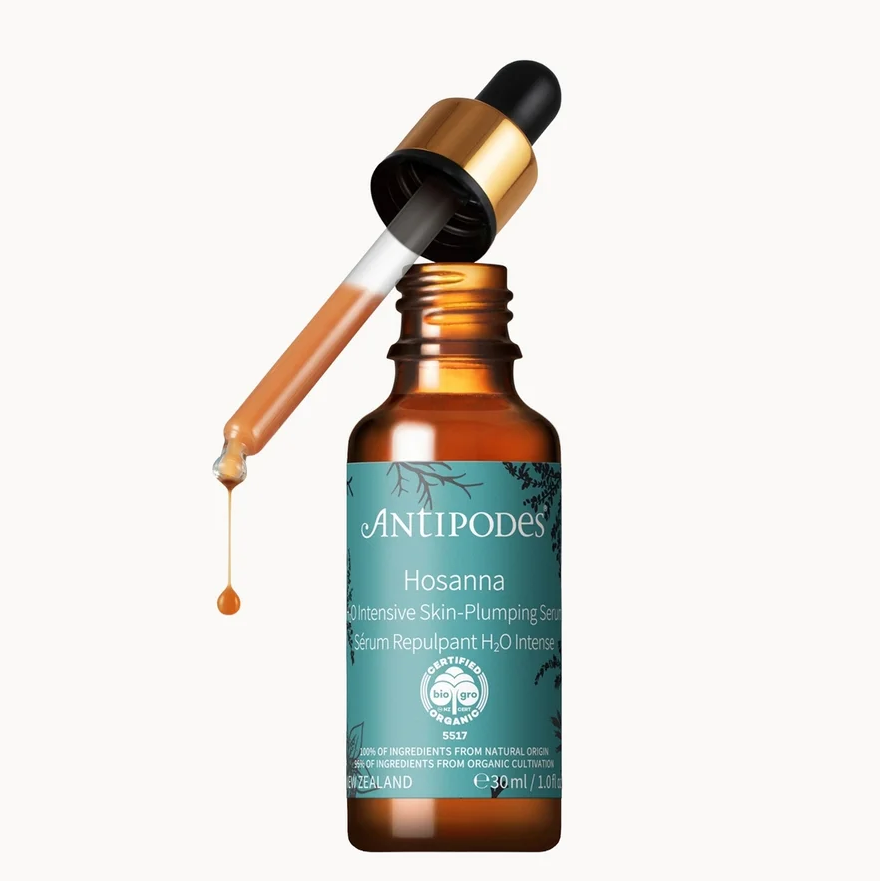 An amber glass dropper bottle of Antipodes Cosmetics Hosanna serum with a teal label, shown with the dropper tilted to release a single drop of liquid against a plain white background.