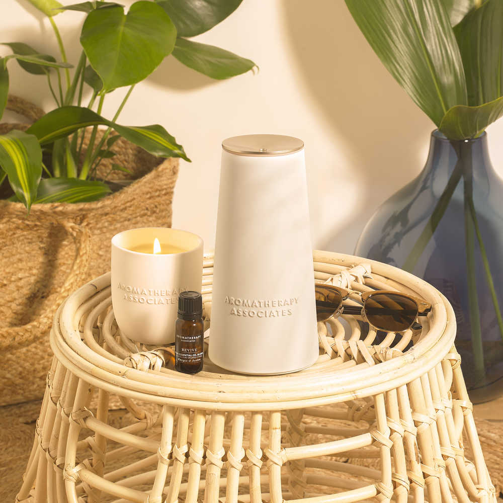 a candle, a diffuser and a bottle of essential oil on a woven table with plants in the background