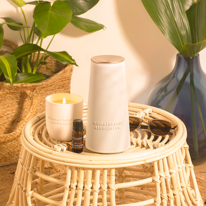 a candle, a diffuser and a bottle of essential oil on a woven table with plants in the background