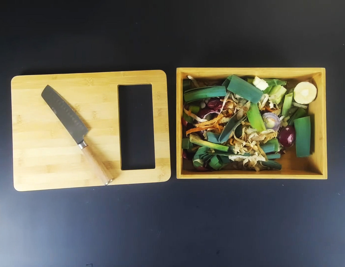 Wooden cutting board with a knife and black stone, next to a wooden box filled with sliced vegetables on a dark background.
