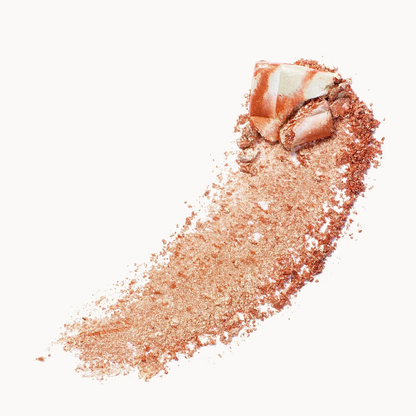 A textured swipe of shimmering rose gold cosmetic powder with a chunky shimmery chunk on top, set against a solid off-white background.