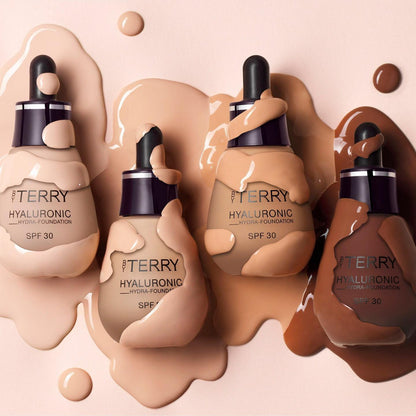 Three dropper bottles of ITERRY Hyaluronic Power Foundation in shades SPF 30, arranged on a peach background with glossy liquid makeup splashes and drips.