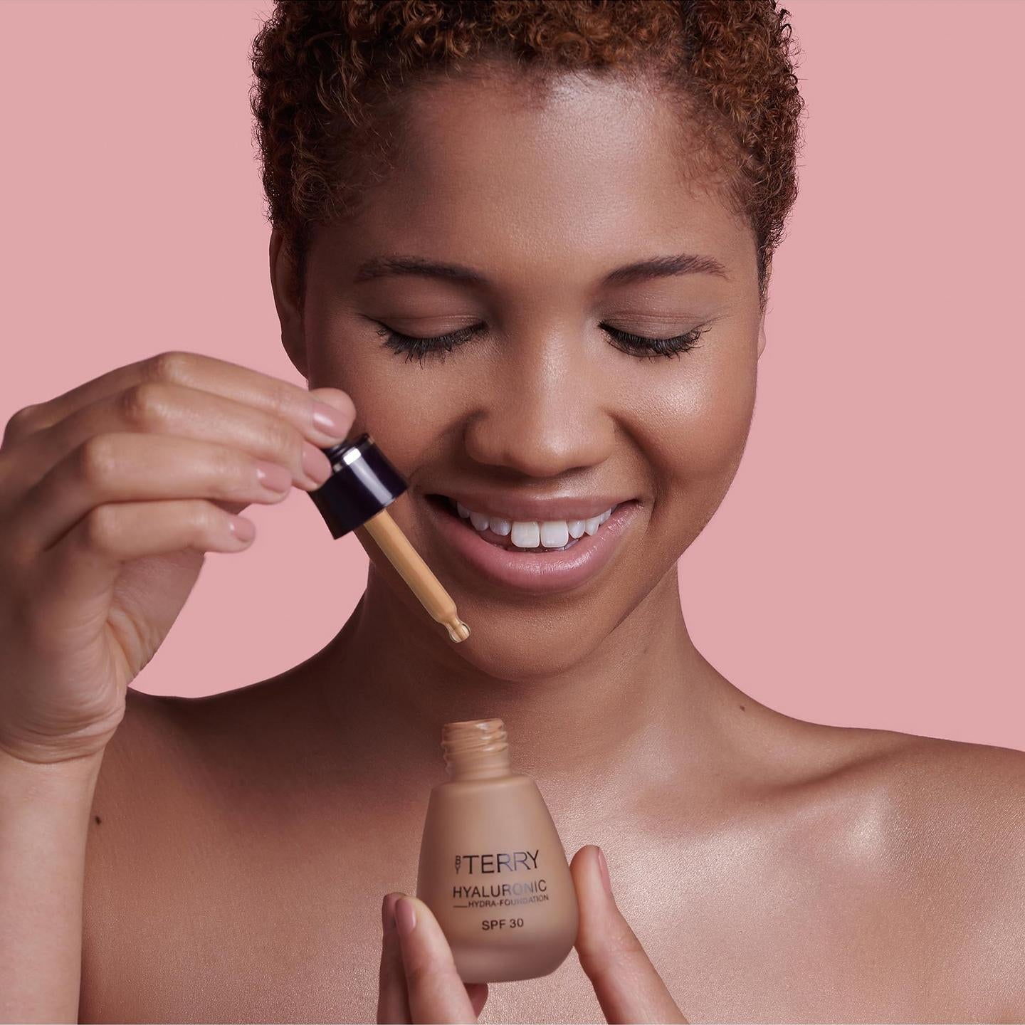 Woman applying foundation with a brush and bottle against a pink background