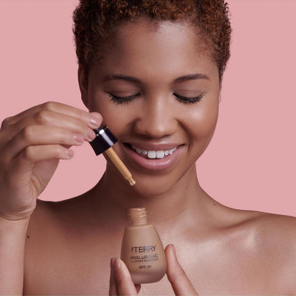 Woman applying foundation with a brush and bottle against a pink background