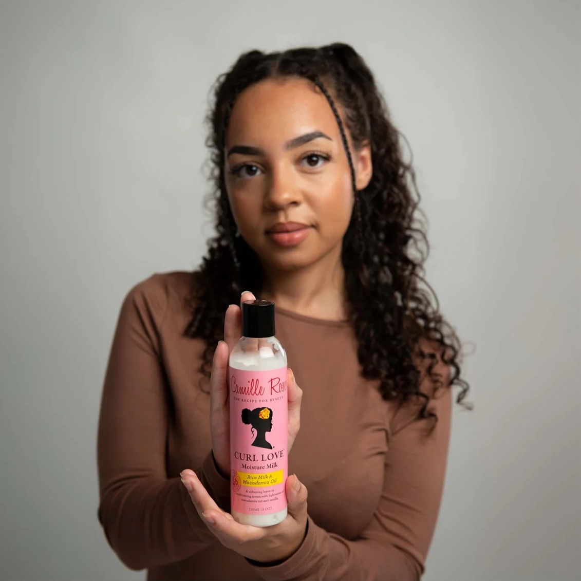 A woman with curly hair wearing a brown long-sleeve shirt holds up a bottle of Curly Love Haircare hair oil against a plain gray studio background.