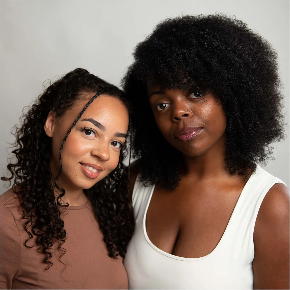 Two women posing together against a light gray studio background. The woman on the right has voluminous dark curly hair and wears a white tank top, while the woman on the left has long dark curly hair and wears a brown top.
