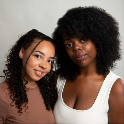 Two women posing together against a light gray studio background. The woman on the right has voluminous dark curly hair and wears a white tank top, while the woman on the left has long dark curly hair and wears a brown top.