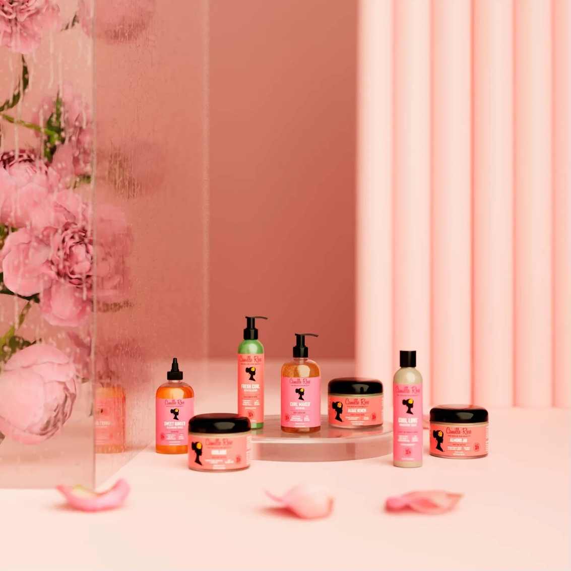 A set of pink haircare and beauty products arranged on a pedestal in a pink studio setting with floral decor and pink petals. The products include pump bottles and jars with black lids, all branded with the same pink label design.