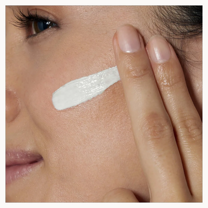 Close-up of a person's face with a thick white cream or balm applied to their cheek and being spread with a finger.