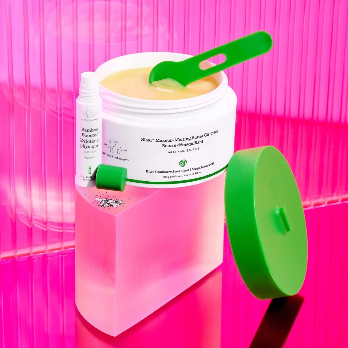 A jar of makeup melting balm with a green scoop inside, shown next to a small bottle and a detached green lid on a bright pink reflective surface.