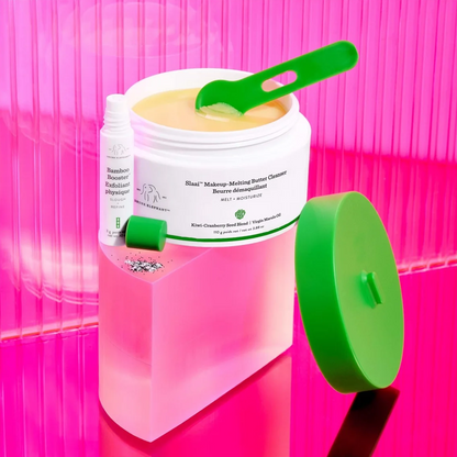 A jar of makeup melting balm with a green scoop inside, shown next to a small bottle and a detached green lid on a bright pink reflective surface.