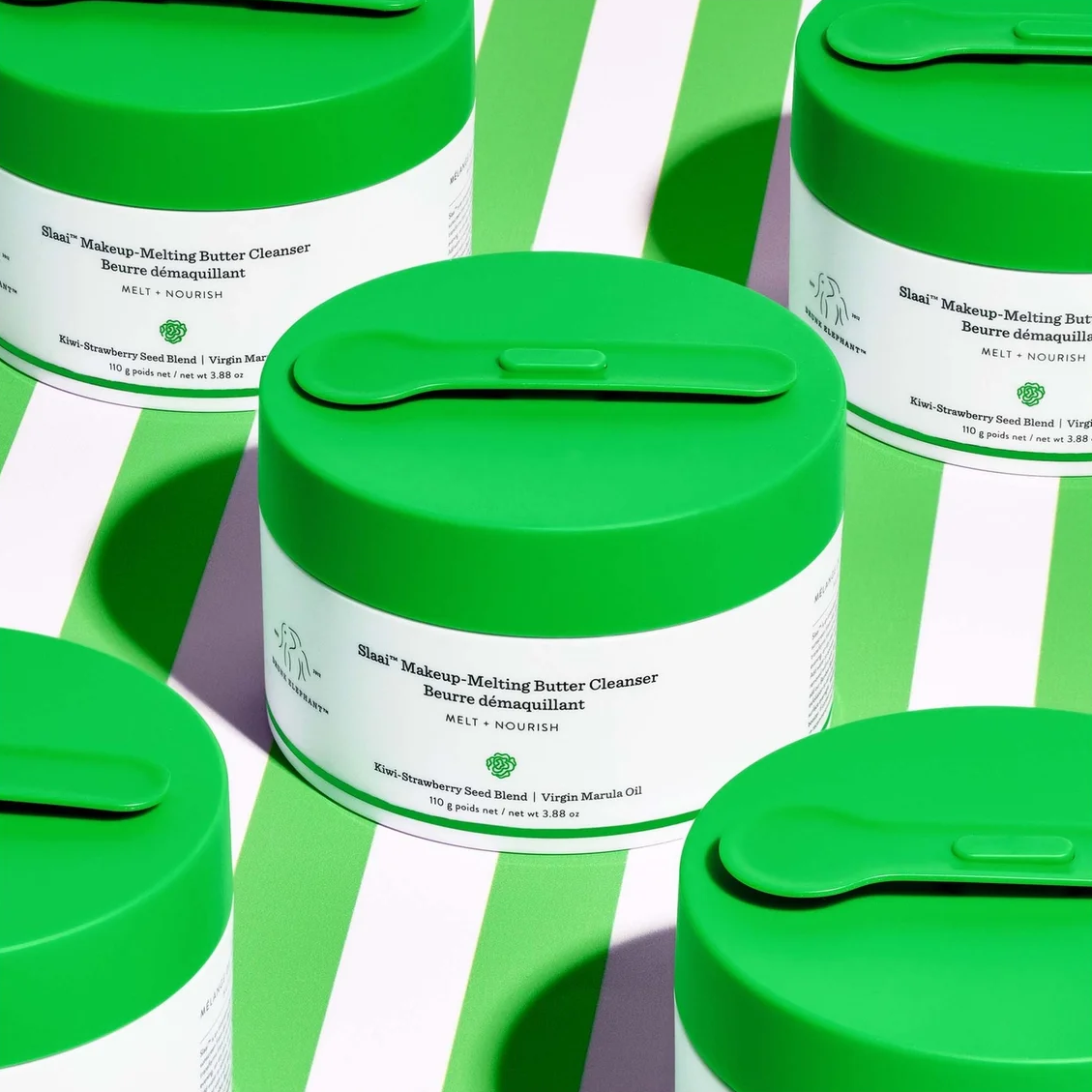 Several white cosmetic jars with bright green lids arranged on a green-and-white striped background. The front jar label reads Bluebells Makeup-Melting Butter Cleanser.