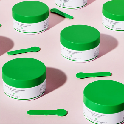 Several white cosmetic jars with bright green lids are arranged on a pale pink background, each accompanied by a small green spatula. The product packaging is clearly visible, though the text is too small to read confidently.