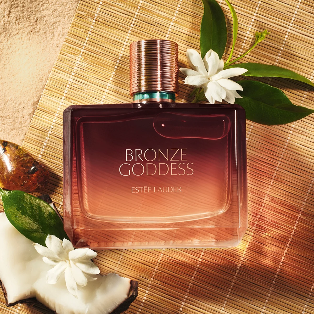 Estee Lauder Bronze Goddess perfume bottle on a textured surface with flowers and leaves.