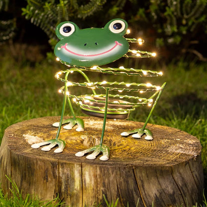 A green metal frog garden light sculpture with glowing warm white wire lights, displayed on a wooden tree stump in an outdoor grassy setting at night.