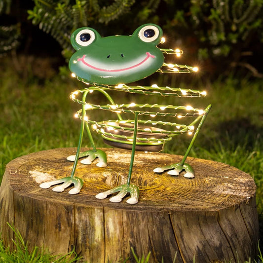 A green metal frog garden light sculpture with glowing warm white wire lights, displayed on a wooden tree stump in an outdoor grassy setting at night.
