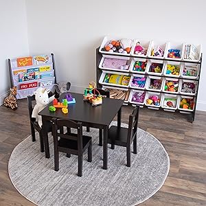 A children's playroom scene with a small black table and four matching chairs on a round gray rug, surrounded by toy bins, a bookshelf, and wood flooring.