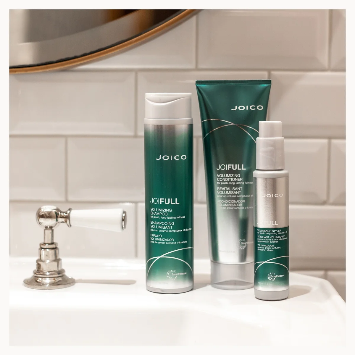 Three Joico JOFULL haircare products in teal and silver packaging are lined up on a white bathroom sink with a tiled wall and mirror in the background.