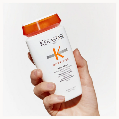A hand holding a white bottle of Kérastase Paris Nutritive Bain Sâin (hair bath) with an orange cap against a clean white background.