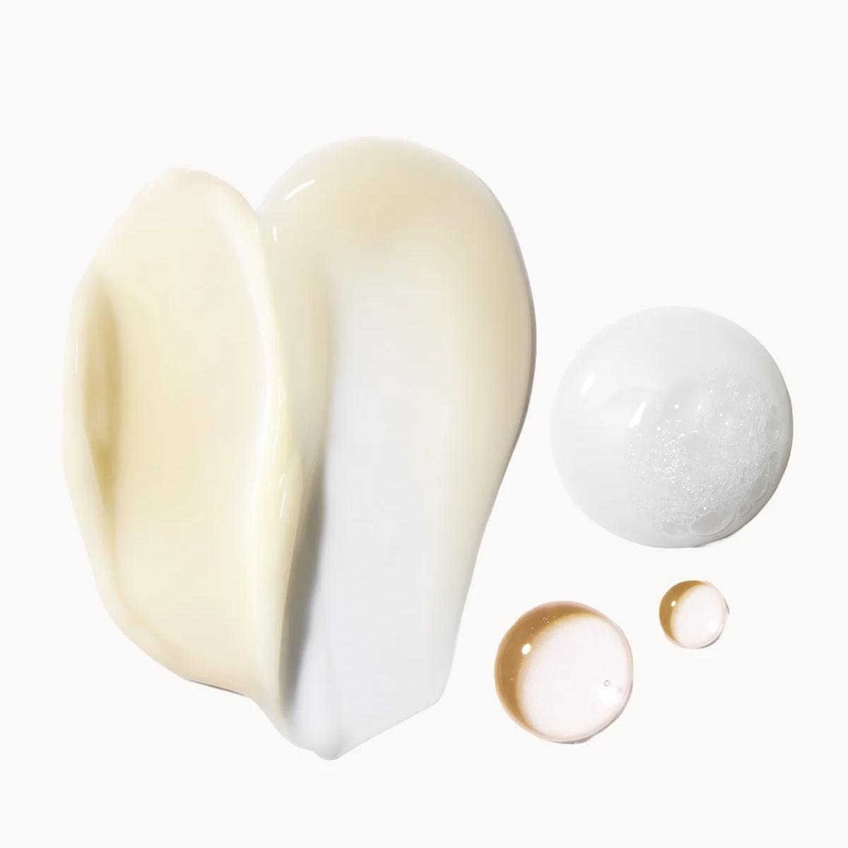 A cosmetic product shown as a large irregular smear of creamy off-white product next to three glossy droplets of the same formula on a plain white background.