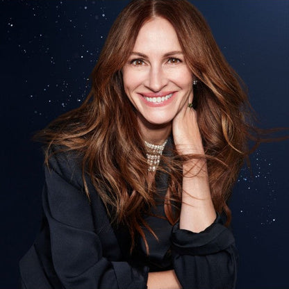 A smiling actress julia roberts with long wavy brown hair, wearing a dark navy top and a pearl necklace, posing against a dark navy blue background with small white speckles.