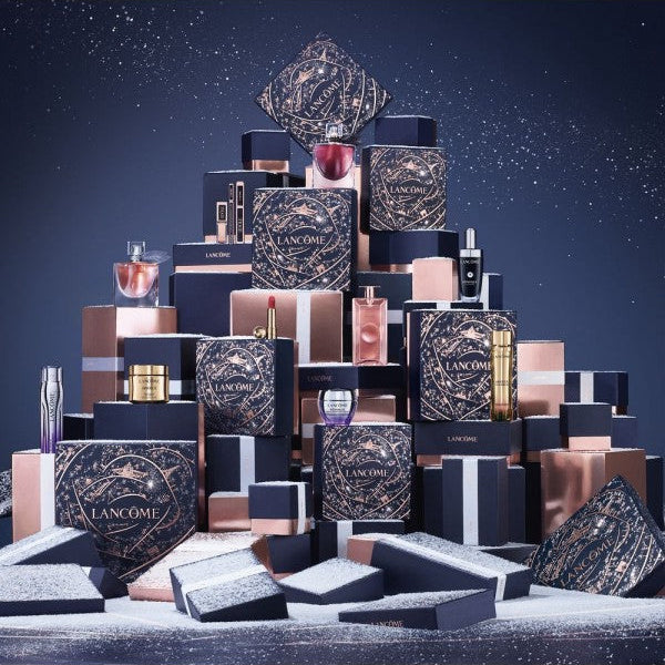 A luxury cosmetic gift set arranged in a pyramid stack, featuring multiple black and rose gold Lancôme boxes, bottles, and jars on a snowy surface against a dark blue background.