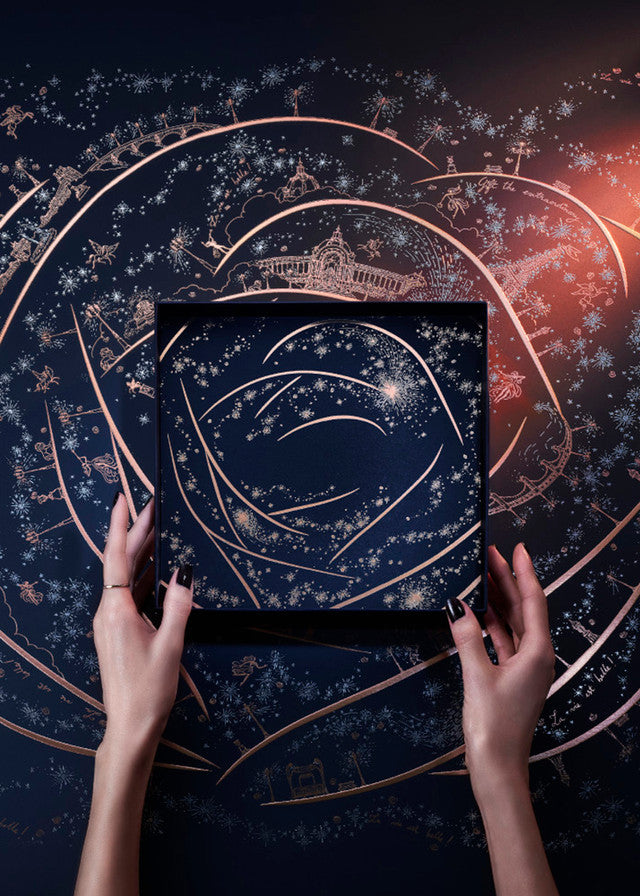 A top-down view of a person's hands holding a square black box decorated with a celestial rose pattern of gold lines and white stars, set against a dark background with rose gold constellations and a warm orange glow.