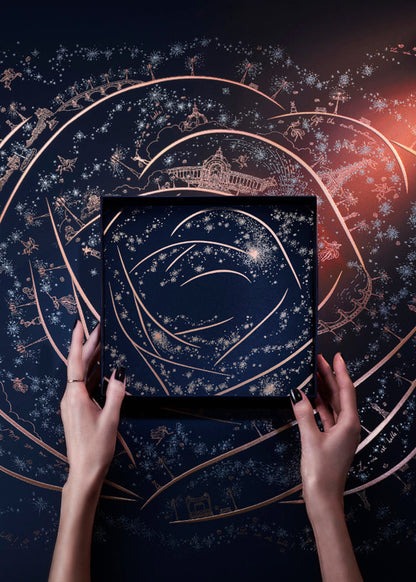A top-down view of a person's hands holding a square black box decorated with a celestial rose pattern of gold lines and white stars, set against a dark background with rose gold constellations and a warm orange glow.