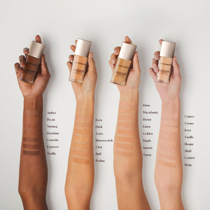 Four hands holding bottles of foundation with swatches on arms against a light background