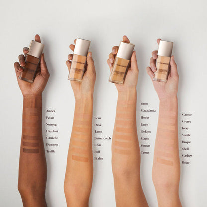 Four hands holding bottles of foundation with swatches on arms against a light background