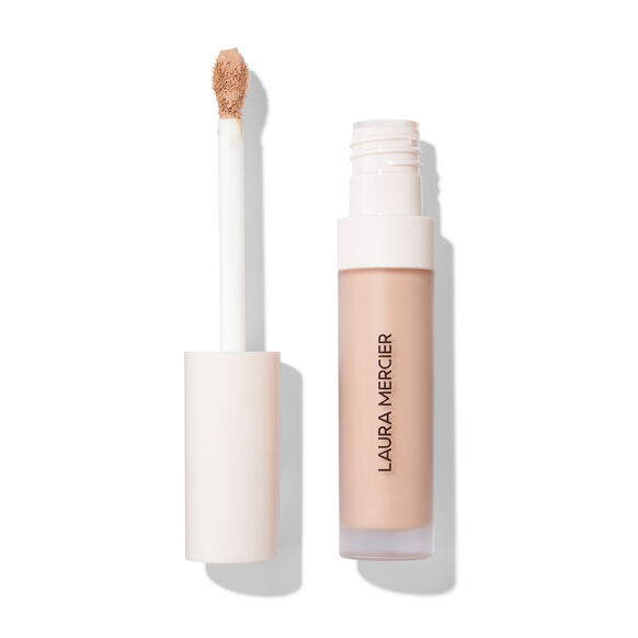 Laura Mercier liquid concealer in a clear tube showing the beige formula, with the cap off and the applicator wand standing beside it on a white background.