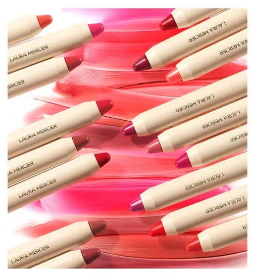Multiple Laura Mercier lip crayons in assorted pink, red, nude, and mauve shades arranged diagonally over a glossy pink and coral painted background.