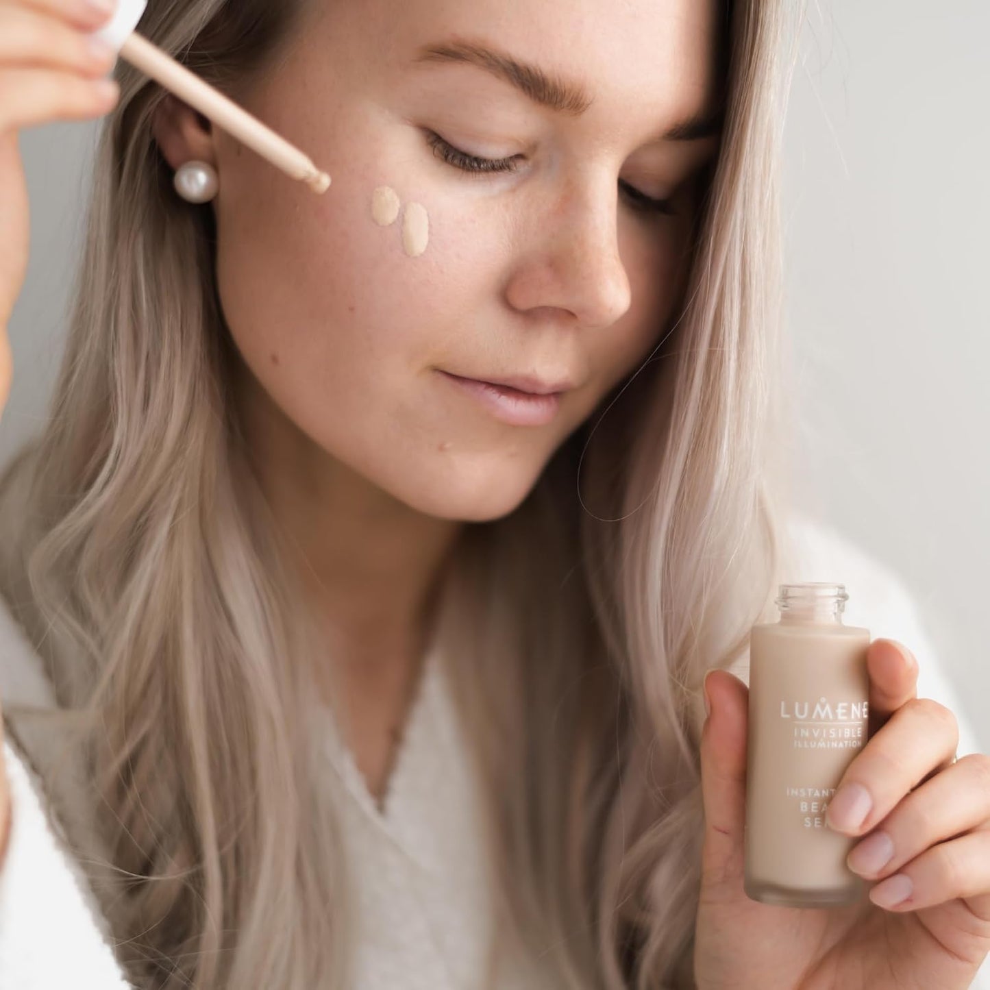 A woman with long blonde hair applies liquid foundation from a dropper to her cheek while holding a bottle of Lumén Invincible Skin Serum foundation. She is wearing a white knit sweater and a pearl earring against a soft, light gray background.