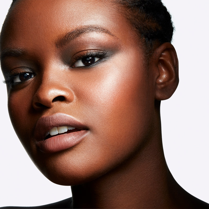 Close-up portrait of a woman with short dark hair, wearing a glossy warm nude lip color and smoky eye makeup against a light gray background.